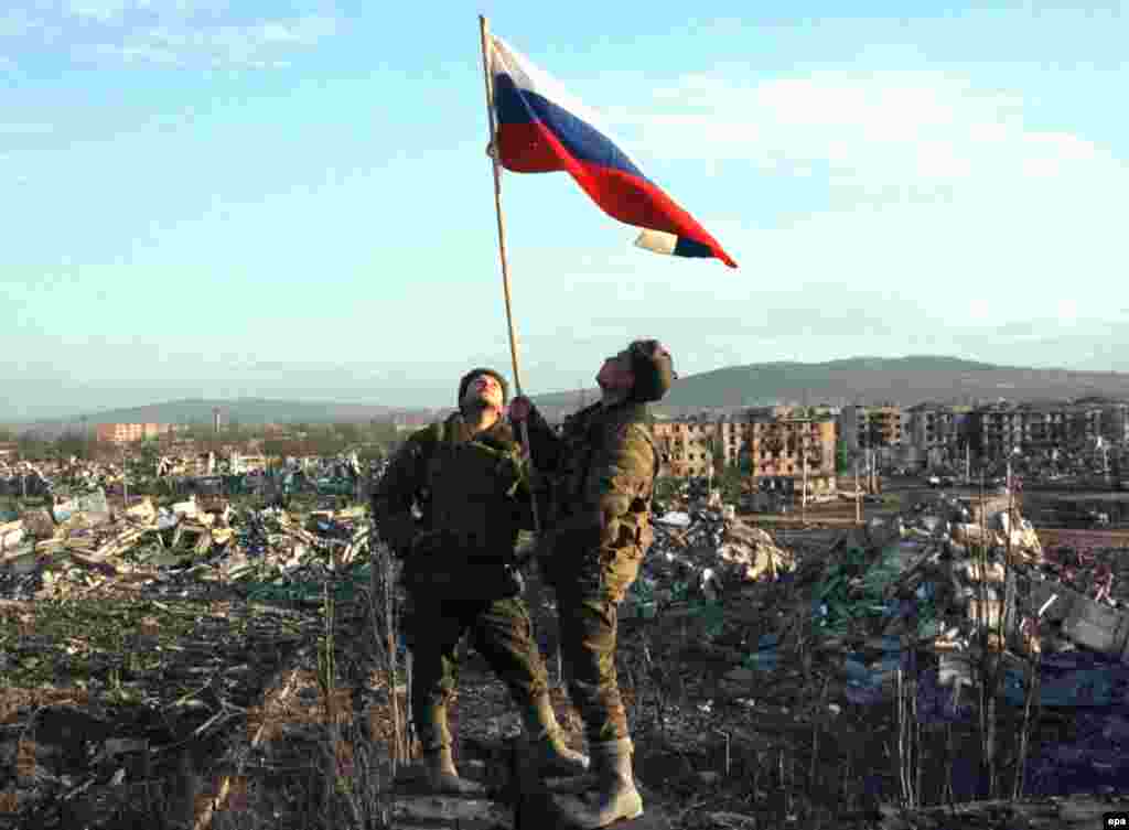 Военные поднимают российский флаг в центре Грозного. Февраль 2000 года. К тому времени, когда федеральные силы установили контроль над городом, большая его часть была превращена в руины после применения бомб и других видов оружия в ходе боевых действий.